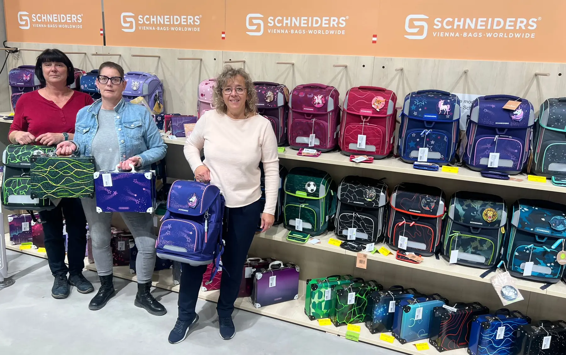 Drei Mitarbeiterinnen zeigen verschiedene Schultaschen vor dem Sortiment